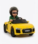 Enfant portant un casque et des lunettes de soleil assis au volant d'une voiture électrique enfant Audi R8 Spyder 12V jaune avec logo quatre anneaux et plaque DLX R8 Spyder